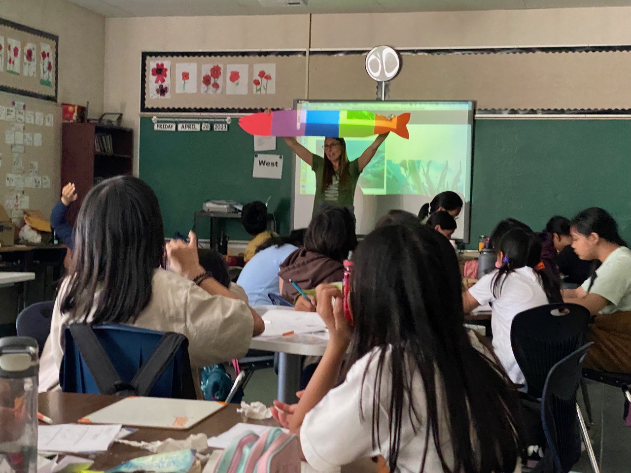 Rowena stands at the front of a classroom, holding a large, colourful paper fish.